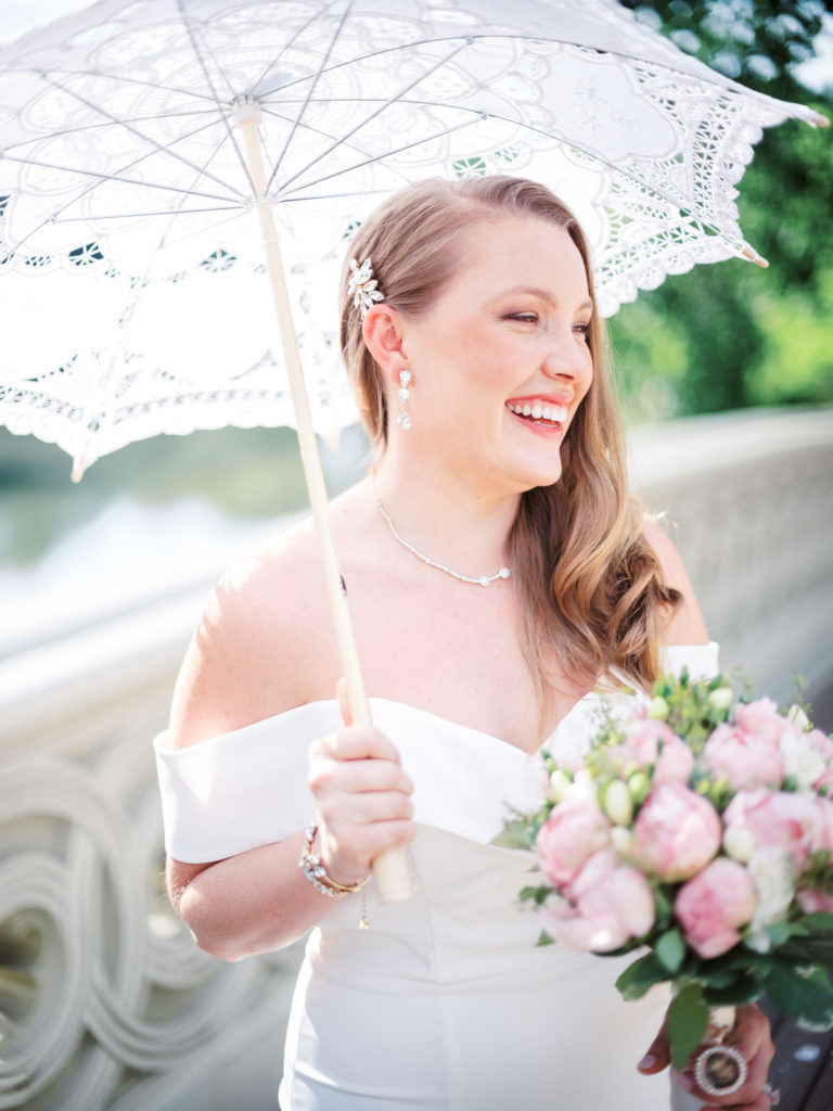 A portrait of the bride wearing a white runway dress with flowers by posies as she stands on the bow bridge the morning of her NYC elopement.