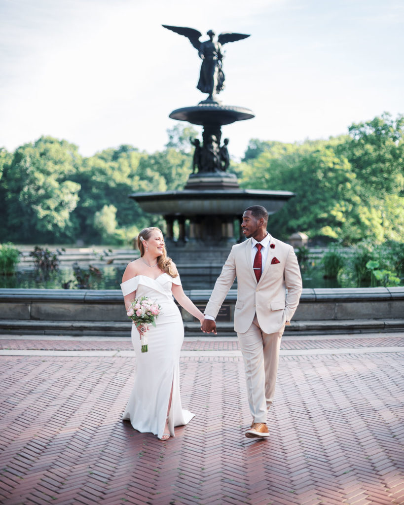 The bride wearing a white runway dress with flower by posies holds hands with her groom in the black tux as they walk together in bethesda terrance and fountain in central park the morning of their NYC elopement.