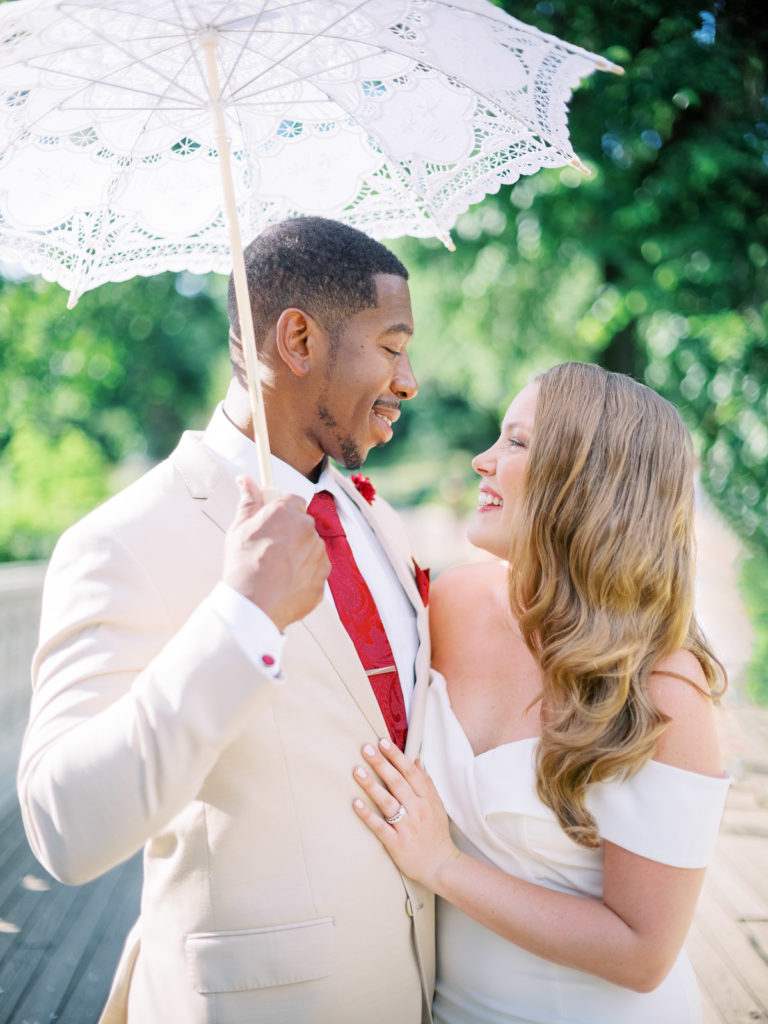 The bride wearing a white runway dress with flower by posies embraces her groom wearing the black tux as they hold an umbrella to block the sun during their NYC elopement.