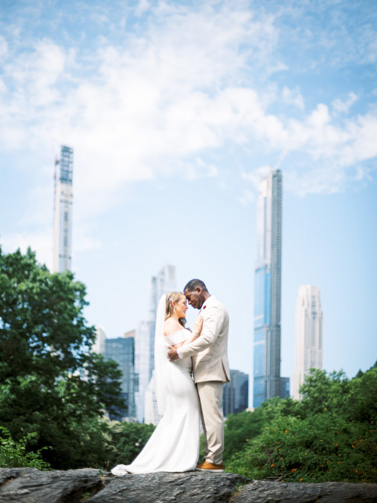 The bride wearing a white runway dress with flower by posies and groom in the black tux pose for a picture atop a hill at the dene summerhouse in central park during their NYC elopement.