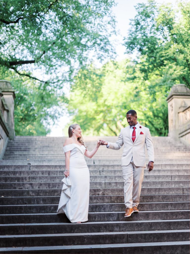 The groom wearing the black tux escorts his bride wearing a white runway dress down the steps at bethesda terrace and fountain in central park on the morning of their NYC elopement.