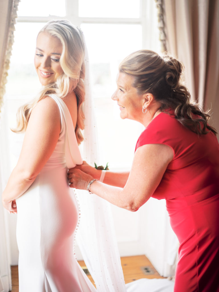 The bride, dressed in Pronovias is being helped into her dress by her mother in one of the first floor rooms in Brecknock Hall before the wedding begins.