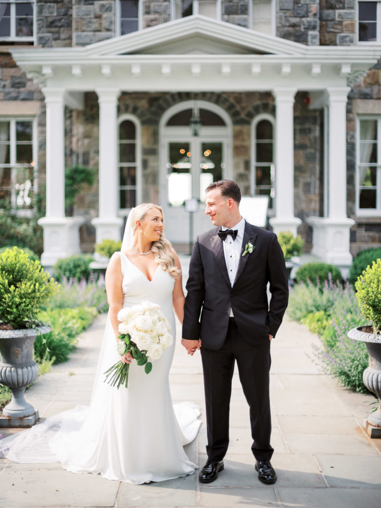 The bride and groom wearing pronovias and The Black Tux with flowers by Kim Jon designs look at each other outside the Mansion during their Brecknock Hall wedding.