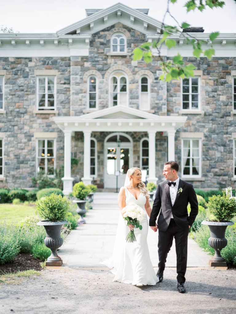 The bride and groom wearing pronovias and The Black Tux with flowers by Kim Jon designs walk outside the mansion at Brecknock Hall.