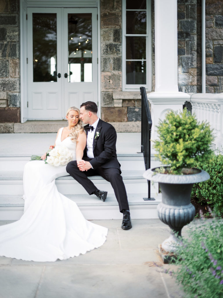 A further away photograph of The bride and groom wearing pronovias and The Black Tux with flowers by Kim Jon designs sitting on the steps outside of the mansion during their Brecknock Hall wedding.