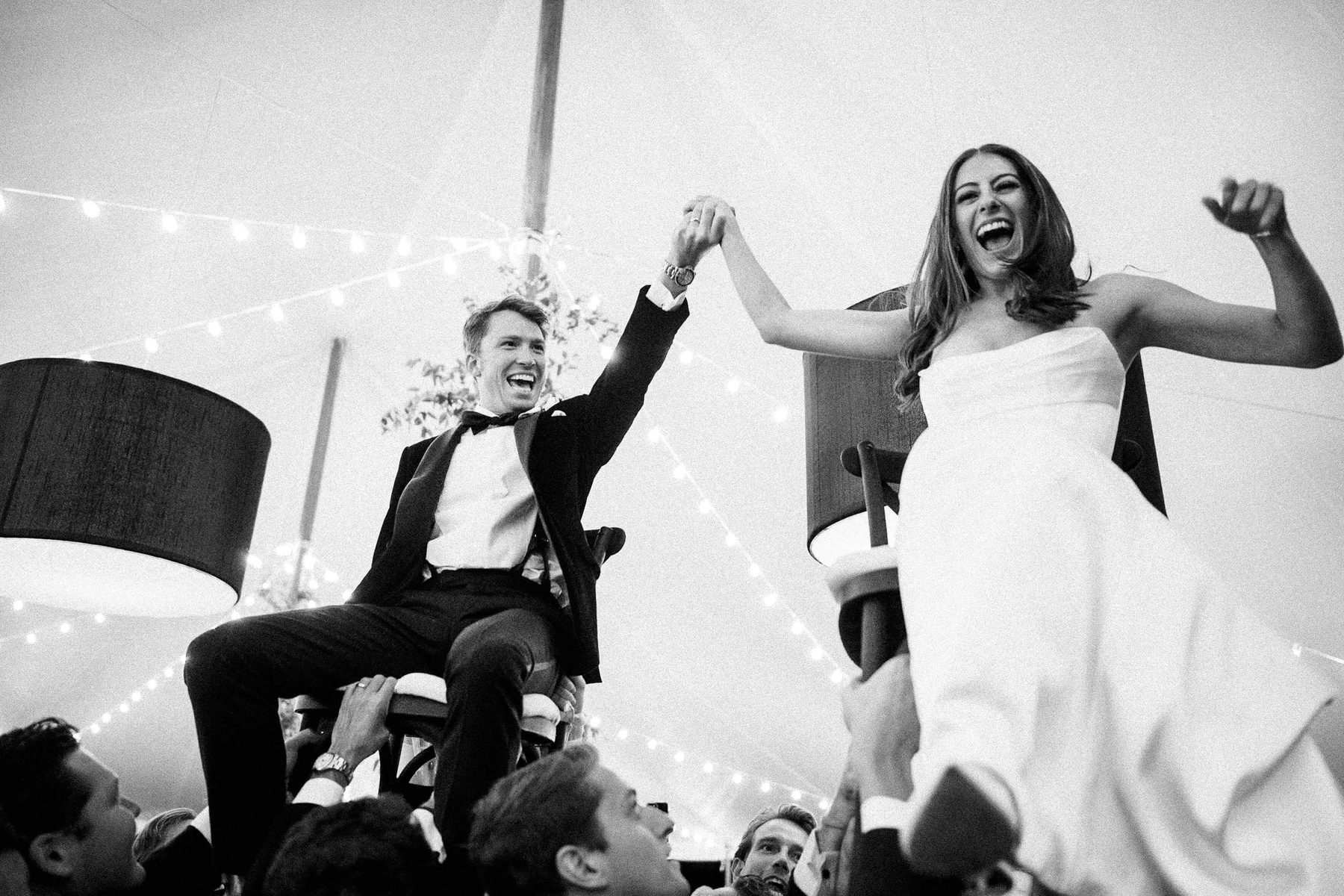 A couple laugh with their hands raised while lifted on chairs during the reception.