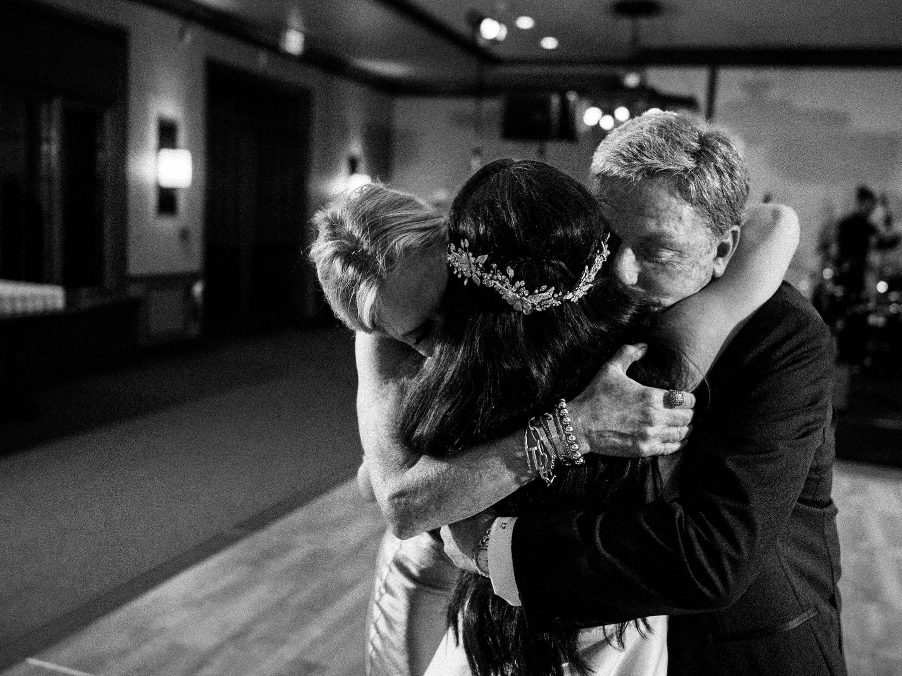 Three people share a close embrace at a wedding, photographed in black and white.