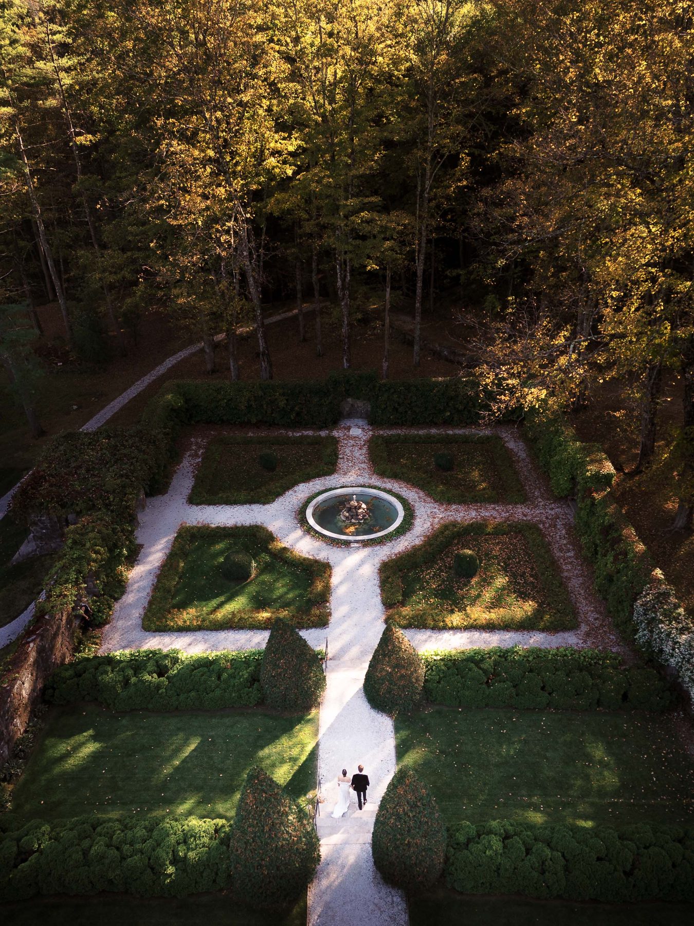 A couple stand in a formal garden framed by trees and paths.